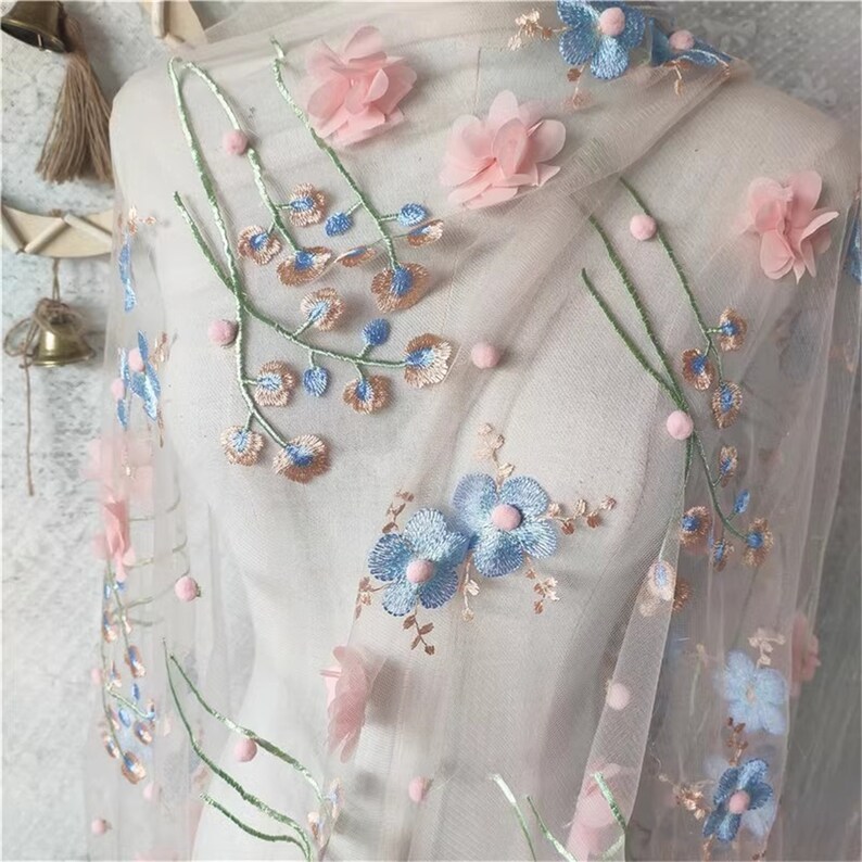 May include: Pale pink tulle fabric with embroidered blue and pink flowers and small white pom-poms.