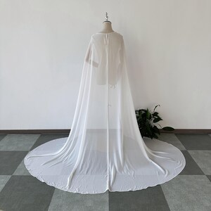 K&ouml;nnte beinhalten: Ein wei&szlig;er, durchsichtiger Chiffon-Umhang mit langem Schleier. Der Umhang ist &uuml;ber einen Mannequin drapiert und hat ein schlichtes, elegantes Design.