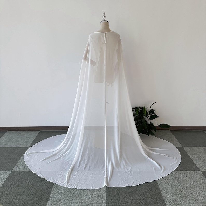 K&ouml;nnte beinhalten: Ein wei&szlig;er, durchsichtiger Chiffon-Umhang mit langem Schleier. Der Umhang ist &uuml;ber einen Mannequin drapiert und hat ein schlichtes, elegantes Design.