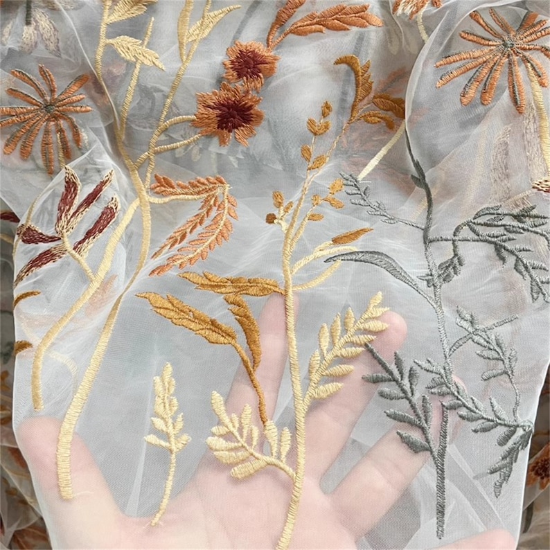 May include: Sheer fabric with embroidered floral design. The embroidery features various botanical elements in shades of brown, gold, and green. The fabric is a light, translucent material.