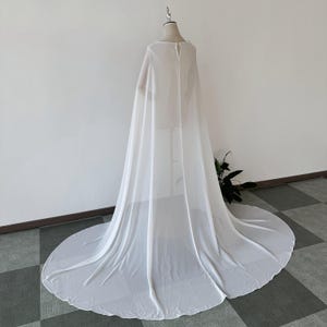 K&ouml;nnte beinhalten: Ein wei&szlig;er, durchsichtiger Chiffon-Umhang mit rundem Ausschnitt und langen &Auml;rmeln. Der Umhang ist &uuml;ber eine Schaufensterpuppe drapiert und hat einen langen Schleier.