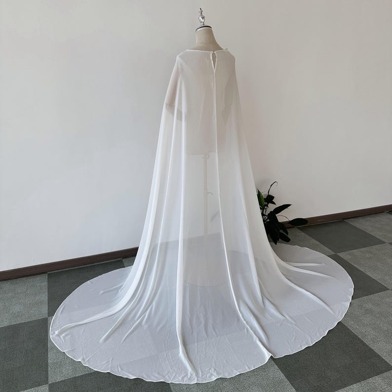 K&ouml;nnte beinhalten: Ein wei&szlig;er, durchsichtiger Chiffon-Umhang mit rundem Ausschnitt und langen &Auml;rmeln. Der Umhang ist &uuml;ber eine Schaufensterpuppe drapiert und hat einen langen Schleier.