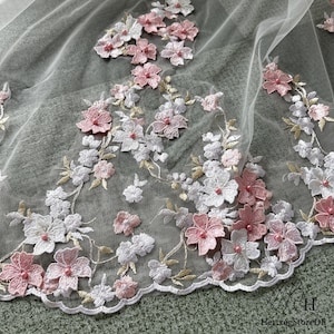 Pu&ograve; includere: Primo piano di un tessuto trasparente e delicato adornato con fiori ricamati. Il design floreale presenta fiori bianchi e rosa con centri perlati, accentuati da steli e foglie color oro. Il bordo del tessuto &egrave; smerlato, creando un aspetto raffinato.