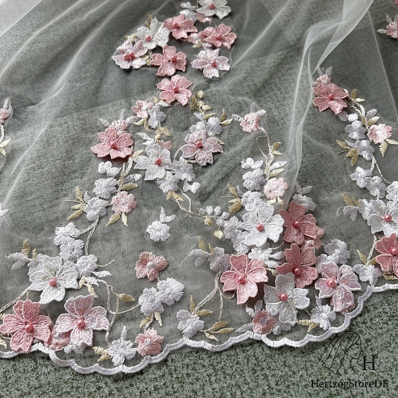 Pu&ograve; includere: Primo piano di un tessuto trasparente e delicato adornato con fiori ricamati. Il design floreale presenta fiori bianchi e rosa con centri perlati, accentuati da steli e foglie color oro. Il bordo del tessuto &egrave; smerlato, creando un aspetto raffinato.