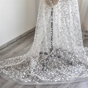 May include: White sheer fabric with an embroidered leaf pattern. The fabric is draped over a surface.