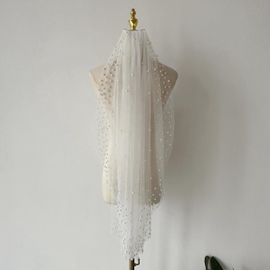 May include: A single-tier tulle bridal veil with pearl embellishments. The veil is displayed on a mannequin.