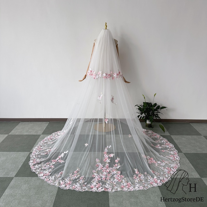May include: A long, white bridal veil with delicate pink floral embroidery along the bottom and a band across the top. The veil is displayed on a mannequin, showcasing its flowing design and intricate details. The veil's train extends gracefully.
