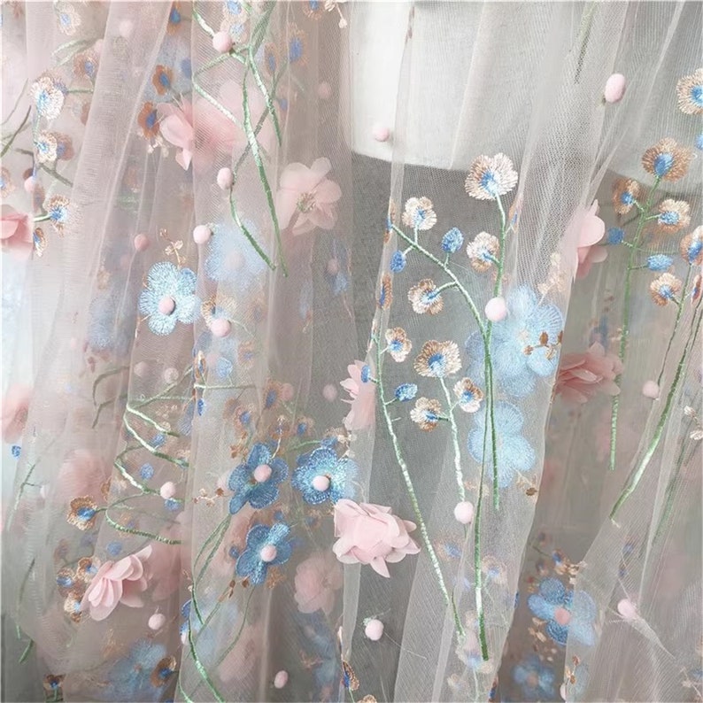 May include: Pink and blue floral embroidered tulle fabric with 3D flowers. The fabric has a delicate, sheer texture and is perfect for crafting and sewing projects.