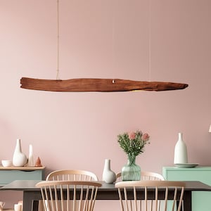 Peut inclure: Une salle à manger avec une table sombre, des chaises en bois clair et un luminaire en forme de poutre en bois. La pièce a un mur rose, un buffet vert et des vases décoratifs avec des fleurs. Les chaises ont un dossier à barreaux.