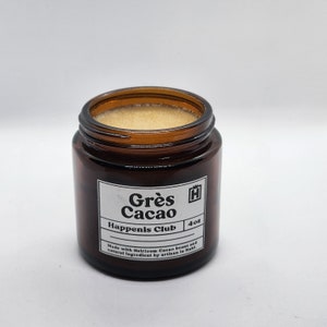 Peut inclure: Un pot en verre brun avec un couvercle dor&eacute; contenant une substance brun clair. Le pot porte une &eacute;tiquette blanche avec du texte noir qui dit "Gr&egrave;s Cacao Happenis Club 4oz Made with Heirloom Cacao beans and natural ingredient by artisan in Haiti."