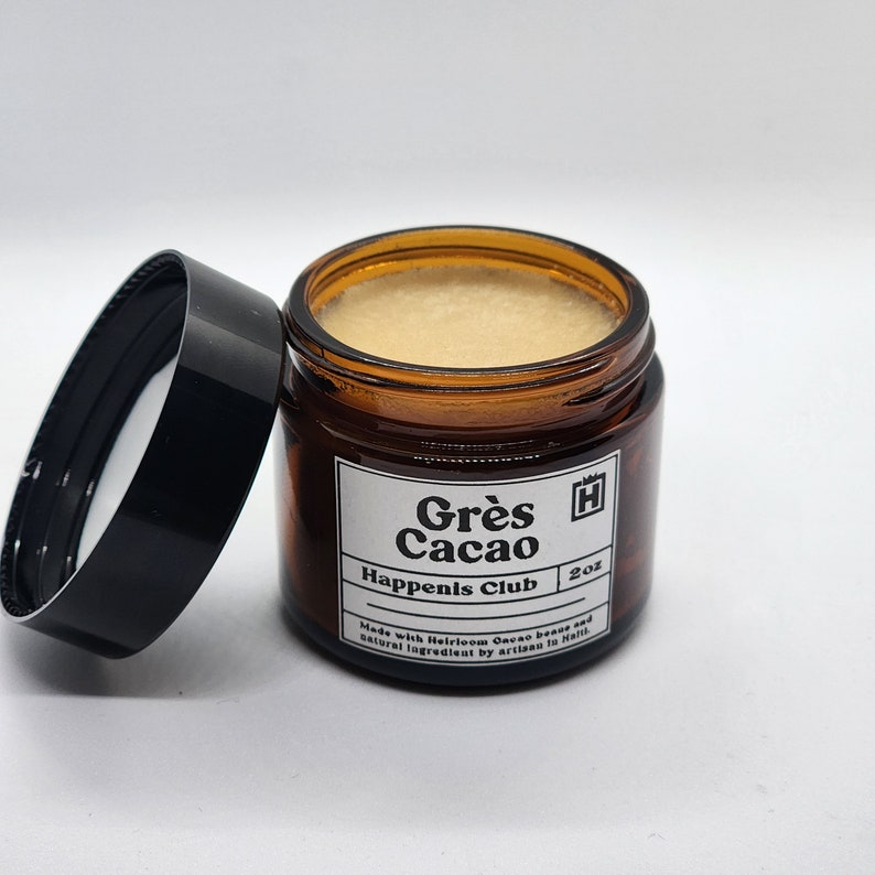 Peut inclure: Un petit pot en verre ambr&eacute; avec un couvercle noir contenant une cr&egrave;me brun clair. Le pot porte une &eacute;tiquette blanche avec du texte noir qui dit "Gr&egrave;s Cacao Happenis Club 2oz Made with Heirloom Cacao beans and natural ingredients by artisans in Haiti."
