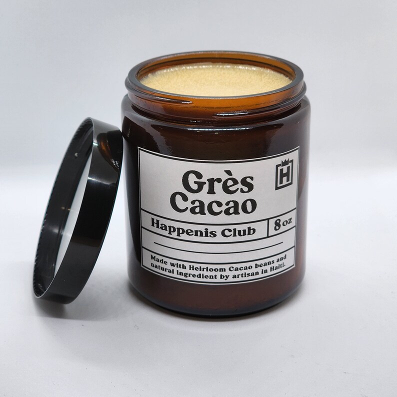 Peut inclure: Un bocal en verre brun avec un couvercle noir contenant une bougie. L'&eacute;tiquette indique "Gr&egrave;s Cacao Happenis Club 8 oz Fabriqu&eacute; avec des f&egrave;ves de cacao h&eacute;ritage et des ingr&eacute;dients naturels par un artisan en Ha&iuml;ti."