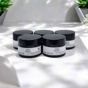 Pode incluir: Cinco potes cosméticos pretos com rótulos brancos são dispostos em grupo. Os rótulos dizem "Grès Cacao" e "Happens Club". Os potes estão sobre uma superfície branca, com sombras. Plantas verdes são visíveis ao fundo.