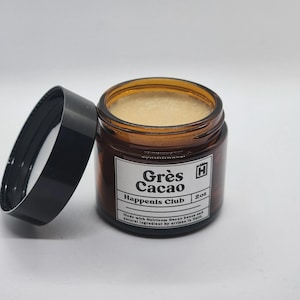 Peut inclure: Un pot en verre ambré avec un couvercle noir contenant une substance beige clair. L'étiquette blanche indique Grès Cacao, Happenis Club, 2oz, et précise qu'il est fabriqué avec des fèves de cacao héritage et des ingrédients naturels en Haïti.