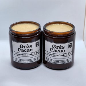 Peut inclure: Deux pots en verre brun avec des &eacute;tiquettes blanches. Les &eacute;tiquettes indiquent "Gr&egrave;s Cacao Happenis Club 8 oz" et "Fabriqu&eacute; avec des f&egrave;ves de cacao anciennes et des ingr&eacute;dients naturels par un artisan en Ha&iuml;ti".