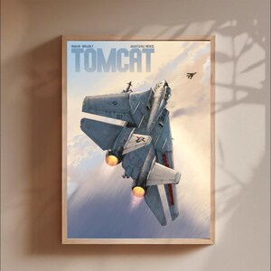 May include: A framed art print featuring a grey F-14 Tomcat fighter jet flying upwards against a cloudy sky. The text TOMCAT is printed in large blue letters at the top, with the names Romain Hugault and Anastasia Reixzl printed above it.