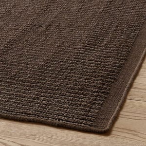 May include: A dark brown rectangular rug featuring a textured surface with horizontal ribbed lines. The rug has a finished stitched border along the edge, showcasing a consistent deep brown color throughout the entire woven fabric construction.