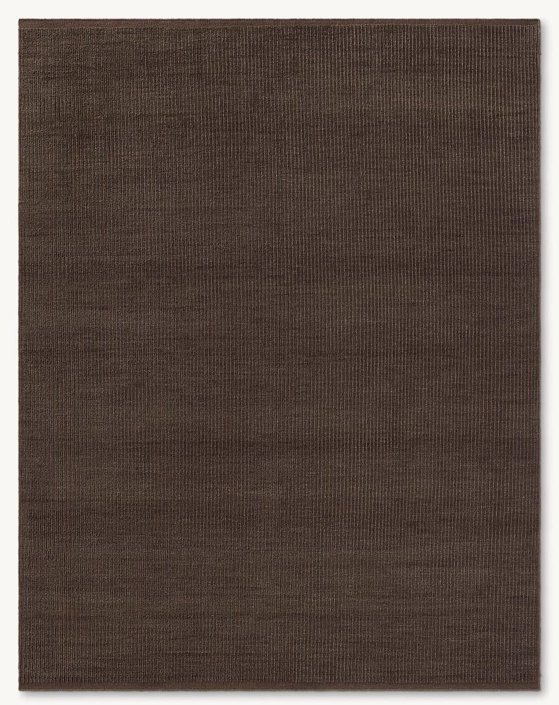 May include: A rectangular area rug in a deep, solid dark brown color. The rug features a consistent, fine vertical ribbed texture across its entire surface, creating a subtle linear pattern. The edges are finished with a clean, matching brown border.