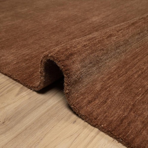 Puede incluir: Primer plano de una alfombra marrón con un borde enrollado. La alfombra tiene una superficie texturizada y está colocada sobre un suelo de madera clara. El color de la alfombra es un tono cálido y terroso.