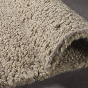 May include: Close-up of a rolled-up, beige, textured rug. The rug has a thick, looped pile, creating a soft, plush appearance. The edges are neatly finished with a contrasting border. The rug is made of a neutral colour.