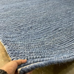 May include: A blue woven rug with a textured surface. The rug is made of thick, braided material and has a light blue colour. The edge of the rug is being held up, revealing a white border.