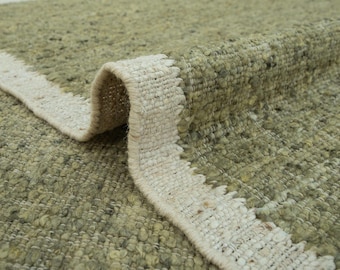 May include: A sage green area rug with a thick, textured, looped pile surface. The rug features a distinct, wide, off-white woven border along the edge where the material folds over, revealing the structural backing and dense, knotted construction.