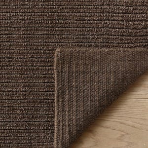 May include: A dark brown area rug with a textured, ribbed surface pattern. The rug is folded over at one corner, revealing the underside of the material, which features a consistent, dense, and woven construction throughout the entire piece.