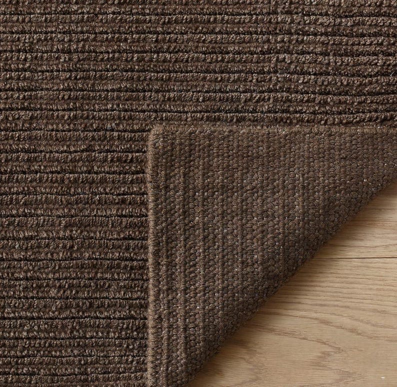May include: A dark brown area rug with a textured, ribbed surface pattern. The rug is folded over at one corner, revealing the underside of the material, which features a consistent, dense, and woven construction throughout the entire piece.