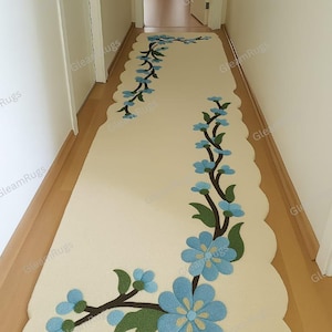 Puede incluir: Una alfombra larga de color crema con borde festoneado y diseño floral. La alfombra presenta un patrón de flores azules y hojas verdes sobre tallos marrones, creando un acento decorativo para el pasillo.