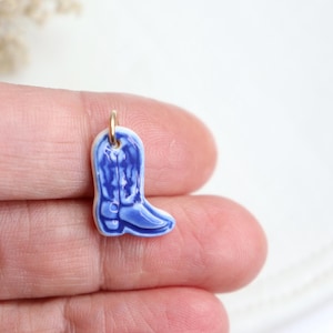 May include: A small, blue ceramic cowboy boot charm with a gold-colored loop for attaching to a necklace or bracelet. The boot has a detailed design and is a miniature representation of a classic Western boot.