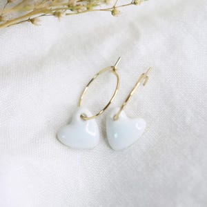 Puede incluir: Un par de pendientes de aro dorados, cada uno con un pequeño dije en forma de corazón blanco, liso y brillante, suspendido de un fino alambre de metal. Los pendientes presentan una estructura circular sencilla y un acabado pulido.