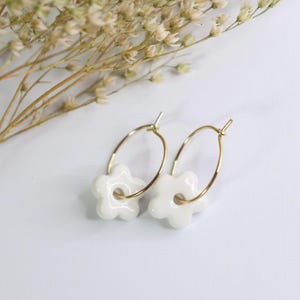 Peut inclure: Une paire de boucles d'oreilles créoles dorées, chacune ornée d'une breloque en forme de fleur blanche à cinq pétales. Les breloques en forme de fleur, lisses et brillantes, sont suspendues à de fins anneaux métalliques dorés.