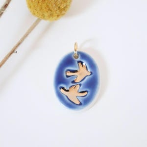 Könnte beinhalten: Ein ovaler Keramikanhänger mit einer tiefblauen, glasierten Oberfläche und zwei eingeprägten, metallisch goldfarbenen Vogelsilhouetten in der Mitte. Der Anhänger verfügt über einen kleinen, goldfarbenen Metallring an der Oberseite.