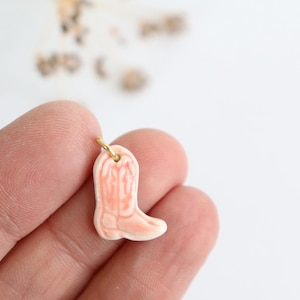 Può includere: Un piccolo ciondolo in ceramica color pesca pallido a forma di stivale da cowboy. Il ciondolo presenta dettagli in rilievo sul gambale e sul piede, con una finitura liscia e lucida. Un piccolo anellino in metallo dorato è fissato in cima.