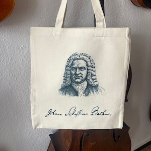 May include: A white canvas tote bag with a black and white illustration of Johann Sebastian Bach. The bag has black handles and the text "Johann Sebastian Bach" is printed in black below the illustration.