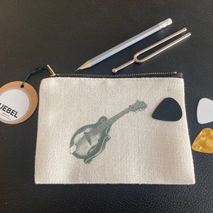 May include: A white canvas zippered pouch with a green and white illustration of a mandolin. The pouch is sitting on a black surface with a black plectrum, a white plectrum, and a gold plectrum next to it.