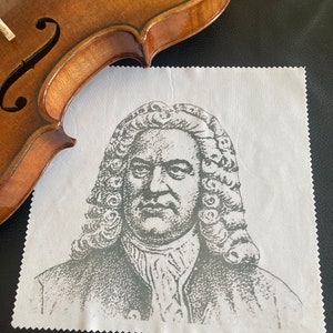 Könnte beinhalten: Ein weißes Mikrofaser-Reinigungstuch mit einer Schwarzweiß-Illustration eines Mannes mit einer langen Perücke und einem ernsten Gesichtsausdruck. Der Mann trägt ein Hemd mit Kragen und eine Jacke. Das Tuch ist in der Mitte gefaltet und hat eine leicht strukturierte Oberfläche.
