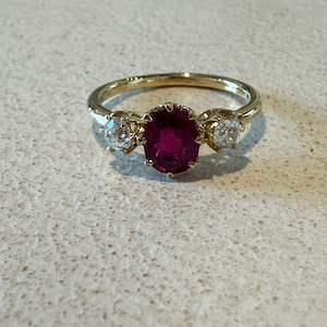 May include: A gold ring featuring a central oval-cut deep red ruby flanked by two smaller round-cut clear white diamonds. The stones are set in a yellow gold band with a prong setting that secures the three gemstones in a horizontal arrangement.