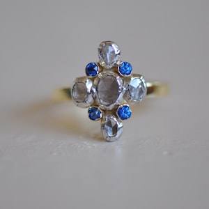 May include: A gold ring with a central cluster of clear, faceted stones and bright blue gemstones. The ring's design is symmetrical, with the stones arranged in a cross-like pattern. The ring is set against a neutral background, highlighting its intricate details.