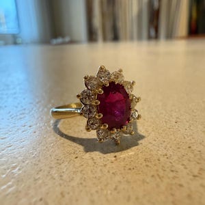 May include: A gold ring featuring a large, round, deep red gemstone surrounded by a halo of smaller, clear gemstones. The ring's band is a simple, polished gold. The setting of the ring is a classic, elegant design.