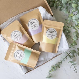 May include: Four small brown paper bags filled with coloured sand. Each bag has a white label with the text "Ceremony Supplies" and the colour of the sand: Pastel Violet, Pastel Pink, Pastel Yellow, and Light Green.