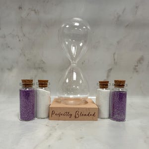 May include: A clear glass hourglass sits on a light-coloured wooden base with the text Perfectly Blended engraved on the front. Four small glass vials with cork stoppers are arranged around the base, two filled with purple sand and two with white sand.