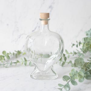 May include: A clear glass bottle shaped like a heart with a narrow neck and a light brown cylindrical cork stopper. The bottle features a smooth, transparent surface and a stable, flat base, designed to hold liquids or decorative contents securely.
