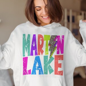 以下が含まれることがあります： 「MARTIN LAKE」とカラフルなグラフィックプリントが施された白いフーディー。中央には稲妻が描かれており、稲妻にはレオパード柄が施されています。