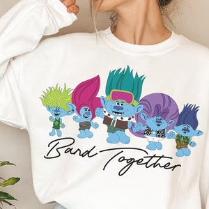 Può includere: Felpa bianca con una grafica di cinque troll blu con i capelli colorati. I troll indossano abiti diversi e hanno espressioni diverse. Il testo "Band Together" è scritto sotto i troll.