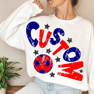 Op de afbeelding: Witte trui met een rode, blauwe en zwarte distressed graphic die "CUSTOM" zegt met een voetbal en sterren.