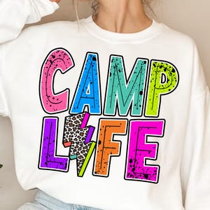 以下が含まれることがあります： 「CAMP LIFE」と雷とヒョウ柄のデザインが施されたカラフルなグラフィックプリントが施された白いスウェットシャツです。