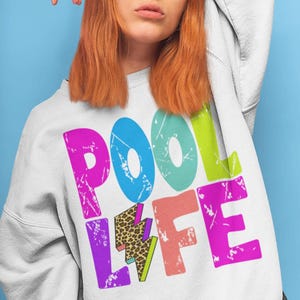 以下が含まれることがあります： 白いスウェットシャツに「POOL LIFE」の文字がカラフルでくたびれた文字で書かれています。文字は黒で縁取られており、80年代のレトロな雰囲気があります。「LIFE」の「L」は、ヒョウ柄の雷です。