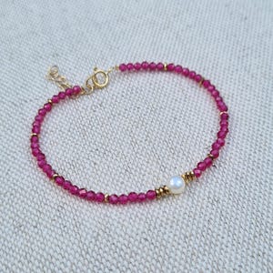 May include: A delicate bracelet made with small pink beads and a single white pearl accent. The bracelet has a gold clasp.