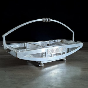 Può includere: Un cestino da portata argentato in stile Art Déco con manico e tre piccole sfere decorative. Il cestino ha una forma rettangolare con un motivo geometrico sul lato e poggia su quattro piedini. Lo sfondo è scuro.